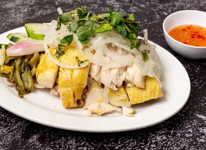 Sliced poached chicken with onions, herbs, pickled vegetables, and a side of chili sauce.