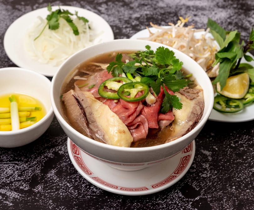 A bowl of pho with beef, cilantro, and jalapeños, surrounded by side dishes of fresh herbs.